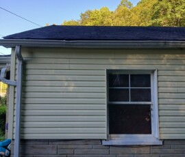 Side view of a house with yellow siding, a single window, and a roof gutter, with trees and blue sky in the background.