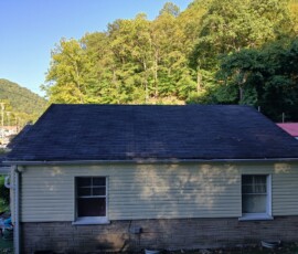 Small yellow house with a black shingle roof, two windows, and trees covering a hillside in the background.