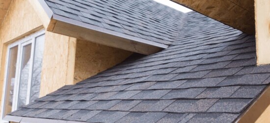 Close-up of a house roof with dark asphalt shingles and unfinished wood siding near a window.