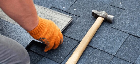 A person wearing an orange glove installs asphalt shingles on a roof next to a hammer.