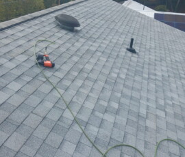 Gray shingled roof with a power drill and extension cord lying near a vent and a pipe, surrounded by trees.