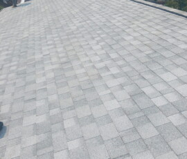 A person in a yellow shirt stands on a large, gray shingle roof under sunlight with trees in the background.
