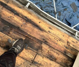 A person stands on worn wooden planks next to a blue tarp and some scattered roofing materials.