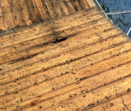 Wooden roof decking with visible water damage and a hole in the center, tarps visible on the ground below.
