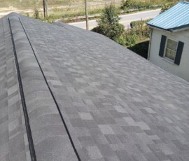 Gray asphalt shingle roof seen from above, with a road, trees, and a blue-roofed house in the background.