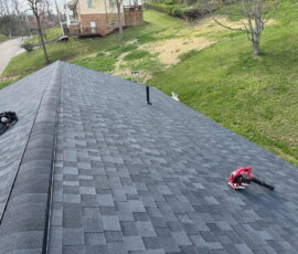 Person using a leaf blower to clean a gray shingle roof on a house with grass and trees in the background.