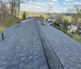 View of a shingled house roof with tools on top, overlooking a suburban neighborhood and distant hills.